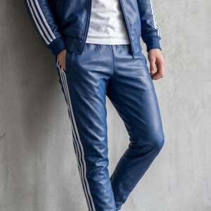 May include: Blue leather tracksuit with white stripes. The jacket has a hood and the trousers have a straight leg. The person is wearing a white t-shirt and white trainers. The outfit is leaning against a grey wall.
