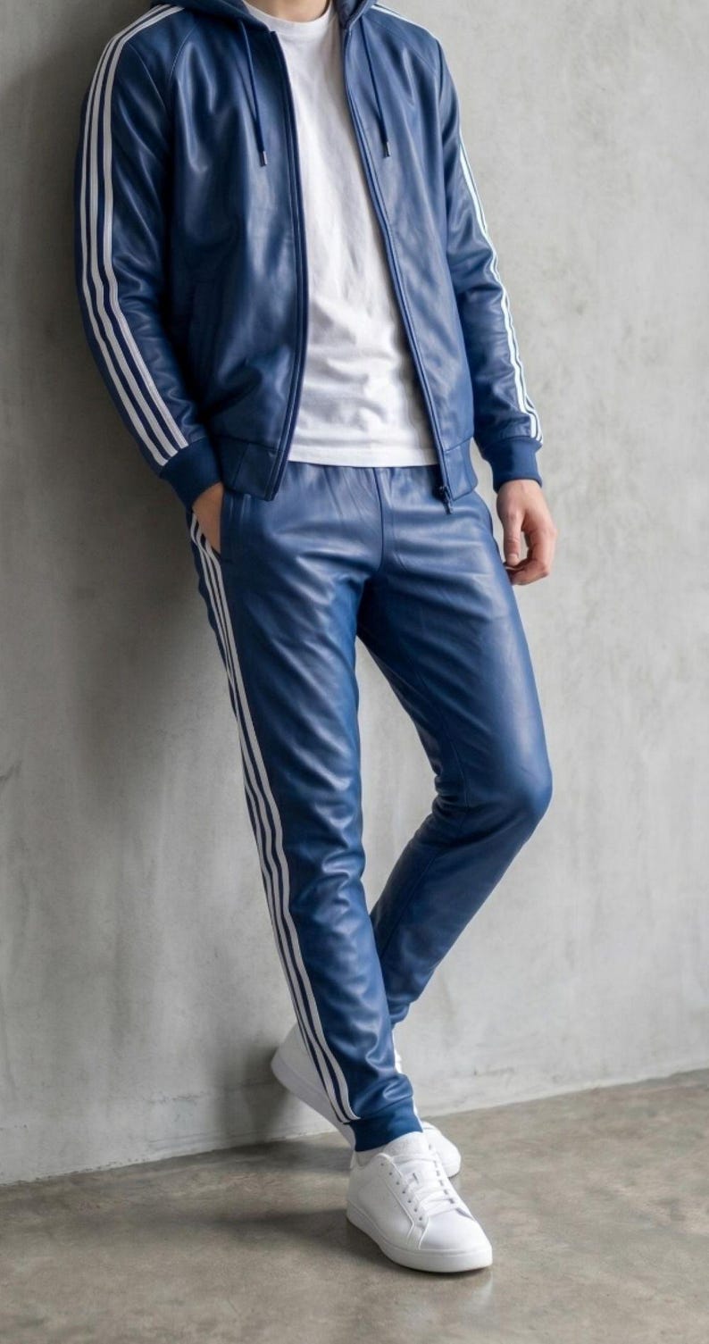 May include: Blue leather tracksuit with white stripes. The jacket has a hood and the trousers have a straight leg. The person is wearing a white t-shirt and white trainers. The outfit is leaning against a grey wall.