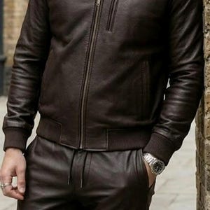 May include: A brown leather bomber jacket with a zip and ribbed cuffs and collar. The jacket is worn with matching leather trousers. The outfit is a classic and stylish look.