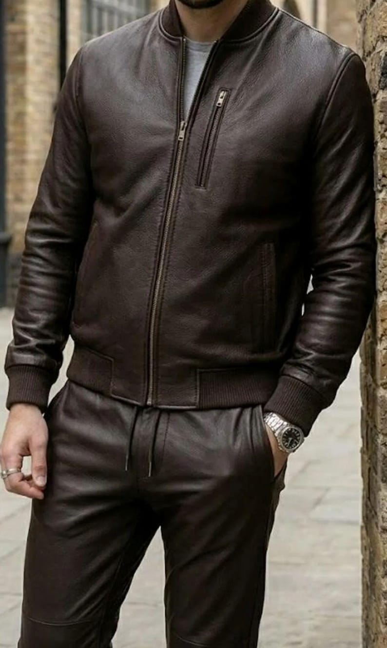 May include: A brown leather bomber jacket with a zip and ribbed cuffs and collar. The jacket is worn with matching leather trousers. The outfit is a classic and stylish look.