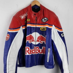 Puede incluir: Una chaqueta de moto de cuero roja, blanca y azul con el logotipo de Red Bull en el pecho. La chaqueta incluye varios parches de patrocinadores como Motul, Arai y NGK, una cremallera frontal completa, un cuello mao y paneles laterales blancos.