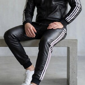 May include: Black leather tracksuit featuring white stripes on the sleeves and legs. The jacket has a zip, and the trousers have a drawstring. White trainers complete the outfit. The person is sitting on a concrete bench.