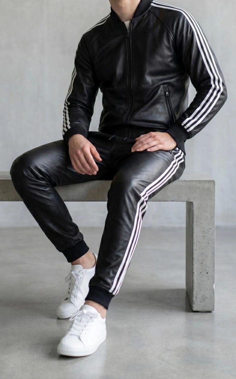 May include: Black leather tracksuit featuring white stripes on the sleeves and legs. The jacket has a zip, and the trousers have a drawstring. White trainers complete the outfit. The person is sitting on a concrete bench.