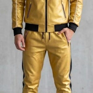 May include: A gold tracksuit featuring a hooded jacket and matching trousers. The jacket has black and white shoulder detailing and a zip. The trousers have a black stripe down the side. The outfit is made of a shiny material, suitable for casual wear.