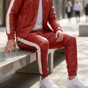 May include: A red leather jacket and trousers set with white side stripes. The jacket features a button closure, and the trousers have a drawstring. White trainers complete the look. The outfit is a striking fashion choice.
