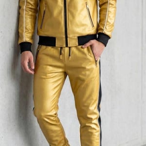 May include: Gold faux leather tracksuit with black and white detailing. The jacket features a hood, zip, and pockets. The trousers have a drawstring waist and a black stripe. The outfit is worn with white trainers.