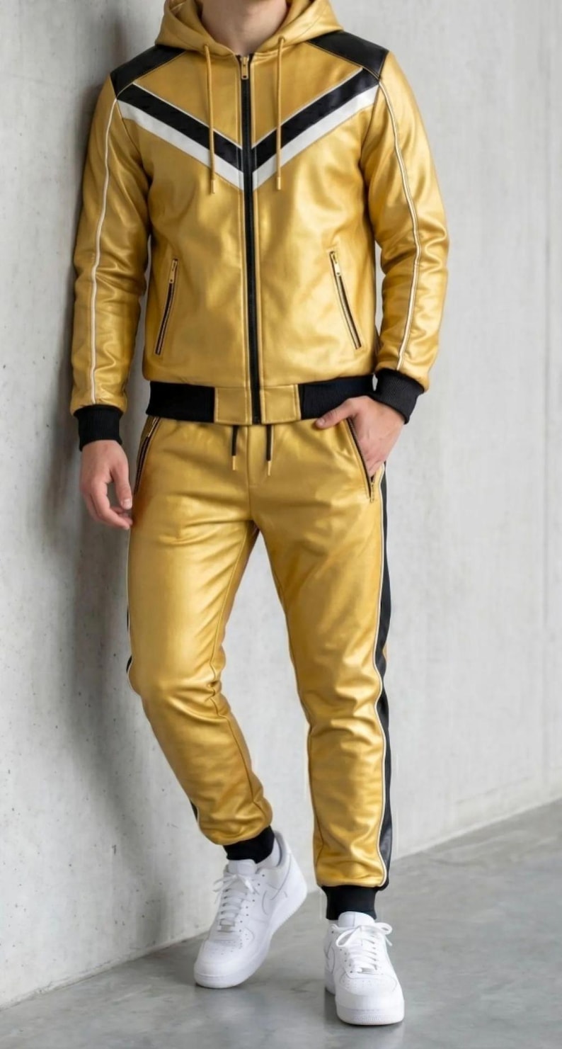 May include: Gold faux leather tracksuit with black and white detailing. The jacket features a hood, zip, and pockets. The trousers have a drawstring waist and a black stripe. The outfit is worn with white trainers.