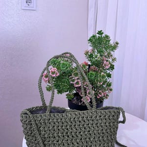 Peut inclure: Un sac fourre-tout vert olive réalisé avec un point de crochet épais et texturé. Le sac présente deux anses tressées robustes et une longue bandoulière assortie, avec une doublure intérieure sombre visible à l'ouverture supérieure du sac.