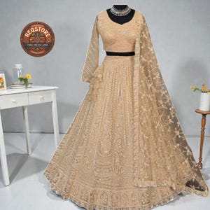 May include: A beige lehenga choli set with intricate embroidery. The set includes a long skirt, a fitted crop top with long sleeves, and a sheer dupatta. The outfit is displayed on a mannequin.