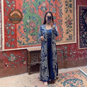 May include: A woman wearing a navy blue embroidered dress with a matching long jacket. The jacket and dress feature intricate silver embroidery. The background includes a vintage rug and the text "REQSTORE AUTHENTIC ETHNIC HERITAGE GOODS SINCE 2020".