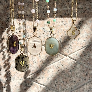 May include: A collection of gold-toned necklaces with gemstone pendants. Each pendant features a different letter, including "C", "A", "W", "Q", and "S". The pendants are in various shapes and colors, with a clover-shaped pendant at the end.