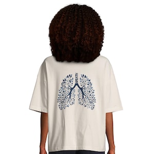 Könnte beinhalten: Cremefarbenes T-Shirt mit einem blau-weißen Lungen-Design auf dem Rücken. Die Lungengrafik besteht aus Blättern und Zweigen, mit dem Wort "BREATH" vertikal darüber gedruckt. Das Shirt hat kurze Ärmel und eine lockere Passform.