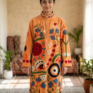 Può includere: Un cappotto arancione a maniche lunghe che arriva al ginocchio, con collo alto e chiusura frontale a zip. Il cappotto è decorato con colorati motivi floreali e a forma di cuore ricamati in blu, rosso, verde e bianco sui pannelli frontali.