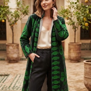 May include: A long, dark green velvet coat featuring vibrant green embroidered pineapple motifs along the front edges and sleeves, worn over a cream-colored camisole and black trousers with a striped inner lining visible.