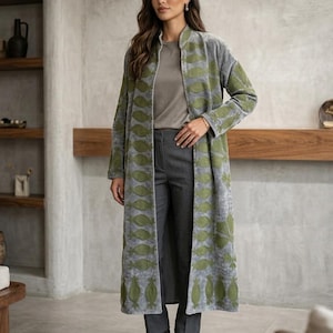 May include: A long, grey, open-front coat featuring a textured, repeating olive green leaf-like pattern along the front panels and sleeves. The coat is worn over a plain grey t-shirt and dark grey trousers, paired with olive green ankle boots.