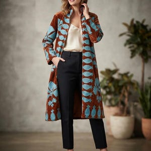 Puede incluir: Un abrigo de largo hasta la rodilla y mangas largas en tela de terciopelo color marrón óxido, con vibrantes patrones bordados en azul claro a lo largo de los bordes frontales y las mangas. Se lleva sobre una camisola crema y pantalones negros.