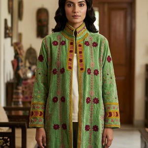 Può includere: Un cappotto a maniche lunghe in velluto verde chiaro con collo alto e apertura frontale. Il capo è decorato con motivi floreali ricamati in rosso scuro e giallo, con dettagli geometrici lungo l'abbottonatura e i polsini.