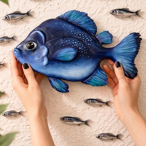 Puede incluir: Una gran escultura de pez azul pintada a mano con detalles en azul claro y motas blancas. Pequeñas esculturas de peces plateados rodean el pez más grande, creando una pieza de arte mural decorativa.