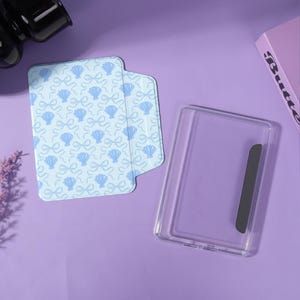 Pode incluir: Uma capa para tablet azul-clara com um padrão repetitivo de laços e conchas brancas, acompanhada por uma capa protetora transparente para tablet que inclui uma faixa magnética cinza-escura no lado direito.