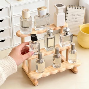 May include: A wooden perfume display stand holding various glass perfume bottles with different cap designs. The stand has two tiers and a wavy edge. The bottles are clear, showcasing the liquid inside.