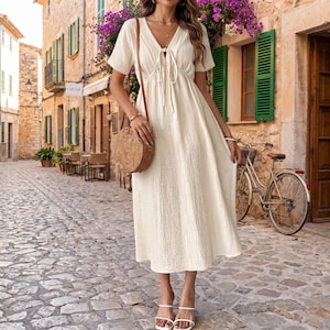 May include: A cream-colored midi dress with short sleeves, a V-neckline featuring a tie-front detail, and a textured fabric finish. The dress is paired with a round, woven straw crossbody bag and white strappy sandals, creating a cohesive look.