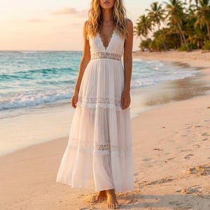 Puede incluir: Un vestido largo blanco con escote en V y detalles de encaje. El vestido tiene una falda escalonada y se usa en una playa con palmeras y el océano de fondo. El vestido está hecho de una tela ligera.