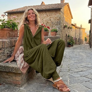May include: An olive green jumpsuit with a button-down front and a relaxed fit. The sleeveless jumpsuit features a hood and pockets. The person is wearing brown sandals and a necklace. The background shows a stone building and a cobblestone street.