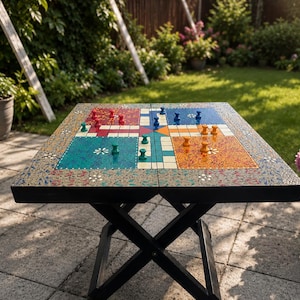 May include: A square folding game table featuring a colorful Ludo board surface with red, blue, teal, and orange quadrants. The table has a black X-shaped base and includes small wooden game pieces in matching colors placed on the board.