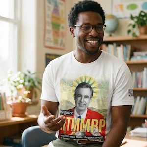 May include: White t-shirt featuring an image of Fred Rogers in a red jacket with the text "He goals: TMMRP would this make Mister Rogers proud?" The image has a yellow sunburst design behind Rogers.