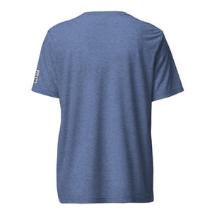 May include: A short-sleeved, heather blue t-shirt. The back of the shirt is visible, with a small, square graphic on the left sleeve. The fabric has a textured appearance. The shirt is a casual wear item.