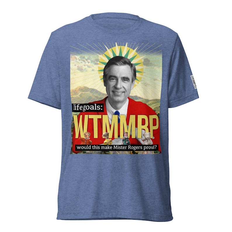May include: Blue heather t-shirt featuring a graphic of Fred Rogers in a red jacket, with the text "life goals: WTMMRP would this make Mister Rogers proud?" The design includes a yellow sunburst and vintage-style illustrations.
