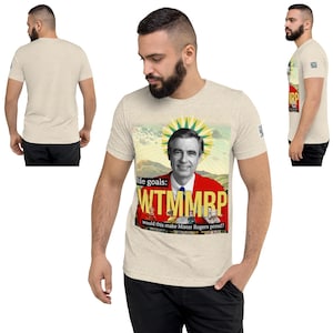 May include: Beige short-sleeve t-shirt featuring a graphic of Fred Rogers in a red jacket with the text "life goals: WTMMRP would this make Mister Rogers proud?" The shirt is made of a soft, comfortable material.