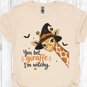 May include: Cream-colored t-shirt featuring a cartoon giraffe wearing a black witch hat with an orange buckle. The text "You bet giraffe I'm witchy" is printed below the giraffe, along with small bat and star graphics. A Halloween-themed graphic tee.