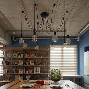 May include: A black, multi-bulb pendant light fitting with exposed Edison-style bulbs hangs from a concrete ceiling. The fixture is above a wooden dining table, creating a modern industrial look. The room features a brick wall and a bookshelf.