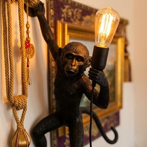 May include: A black monkey-shaped lamp with a vintage-style light bulb. The monkey is holding the bulb and cord, with its tail curled. The lamp is mounted on a wall, with decorative gold rope and a framed picture in the background.