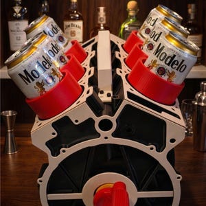 May include: The Pit Stop - Handcrafted V6 Engine Block Can Cooler & Dispenser. A black and white V6 engine block structure with six bright red circular holders containing Modelo beer cans. Includes a red knob at the base and white handle.