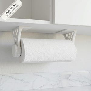 May include: A white under-cabinet paper towel holder with a geometric cutout design on the side brackets and a circular knob on the side. The holder is mounted beneath a white cabinet and holds a roll of white paper towels. Digital STL file.