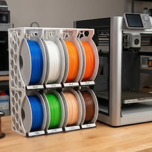 May include: A white 3D printed storage rack with a honeycomb side pattern holding eight circular spools of PLA filament in blue, white, orange, green, tan, and brown. The rack is positioned next to a silver 3D printer with a digital display screen.