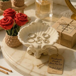 Puede incluir: Un plato decorativo en forma de concha contiene varios anillos. Una pequeña caja de regalo marrón con una etiqueta "Gift Wrap Ready" está cerca. Una etiqueta dice "Handmade Crochet Rose Pot Set Perfect Gift! Etsy". También se ven rosas de ganchillo en una maceta pequeña.