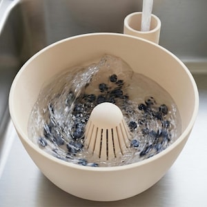 Peut inclure: Un saladier beige pour laver les fruits et légumes, avec une passoire centrale, rempli d'eau et de myrtilles. L'eau coule d'un robinet au-dessus, créant un mouvement tourbillonnant. Le saladier est posé dans un évier en acier inoxydable.