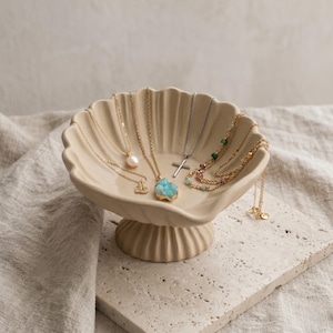 Könnte beinhalten: Eine Sammlung von Halsketten, die in einer beigefarbenen, muschelförmigen Schale präsentiert werden. Die Halsketten weisen verschiedene Anhänger auf, darunter eine Perle, ein Kreuz und ein blauer Edelstein. Die Schale steht auf einer strukturierten, cremefarbenen Oberfläche.
