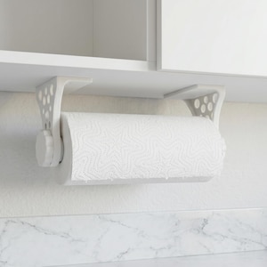 May include: A white, under-cabinet paper towel holder. The holder is mounted under a white cabinet and features a roll of white paper towels. The holder has a decorative, perforated design on the sides and a knob to secure the paper towel roll.