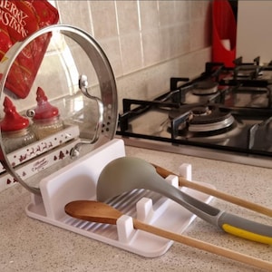 May include: A white kitchen utensil organizer holding a clear glass lid, wooden spoons, and a gray and yellow spatula. A red Christmas-themed oven mitt and jars are visible in the background. The organizer is on a countertop near a gas stove.