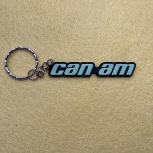 Può includere: Un portachiavi con la scritta "can am" in azzurro, bordata di nero. Il portachiavi è attaccato a un anello e una catena di metallo argentato.