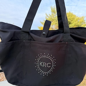 May include: Black tote bag with two long shoulder straps and a small handle. The bag features a white circular design with the letters "KRC" in the center. The bag has side pockets and a snap closure.