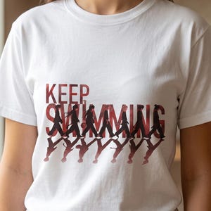 May include: White t-shirt with the words "KEEP SWIMMING" in red. The word "SWIMMING" features a silhouette of people walking. The shirt is a crew neck and has short sleeves.