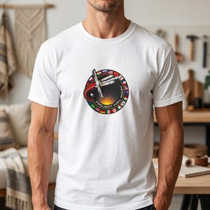 Puede incluir: Camiseta blanca con un diseño circular que representa un transbordador espacial, el texto "PROJECT HAIL MARY" y banderas de varios países. El diseño incluye un arco rojo y naranja, un sol y un borde negro.