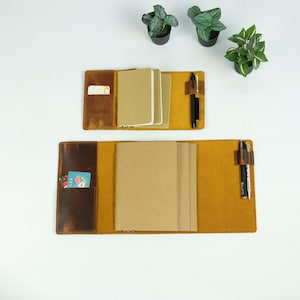 May include: Two open leather notebook covers in tan and dark brown, each featuring internal pockets for cards, multiple beige paper inserts, and a side pen loop holding black pens. The smaller cover has the name Sophia embossed on the left pocket.