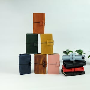 May include: A collection of leather journals in various colors, including brown, green, yellow, blue, and pink. Each journal has a strap closure. A stack of journals in blue, black, and red are also visible.