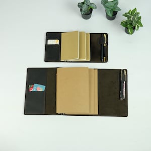 May include: Two dark brown leather notebook covers with internal pockets, each holding multiple tan paper notebooks. The covers feature integrated pen loops containing black and gold pens, and side slots containing small rectangular cards.